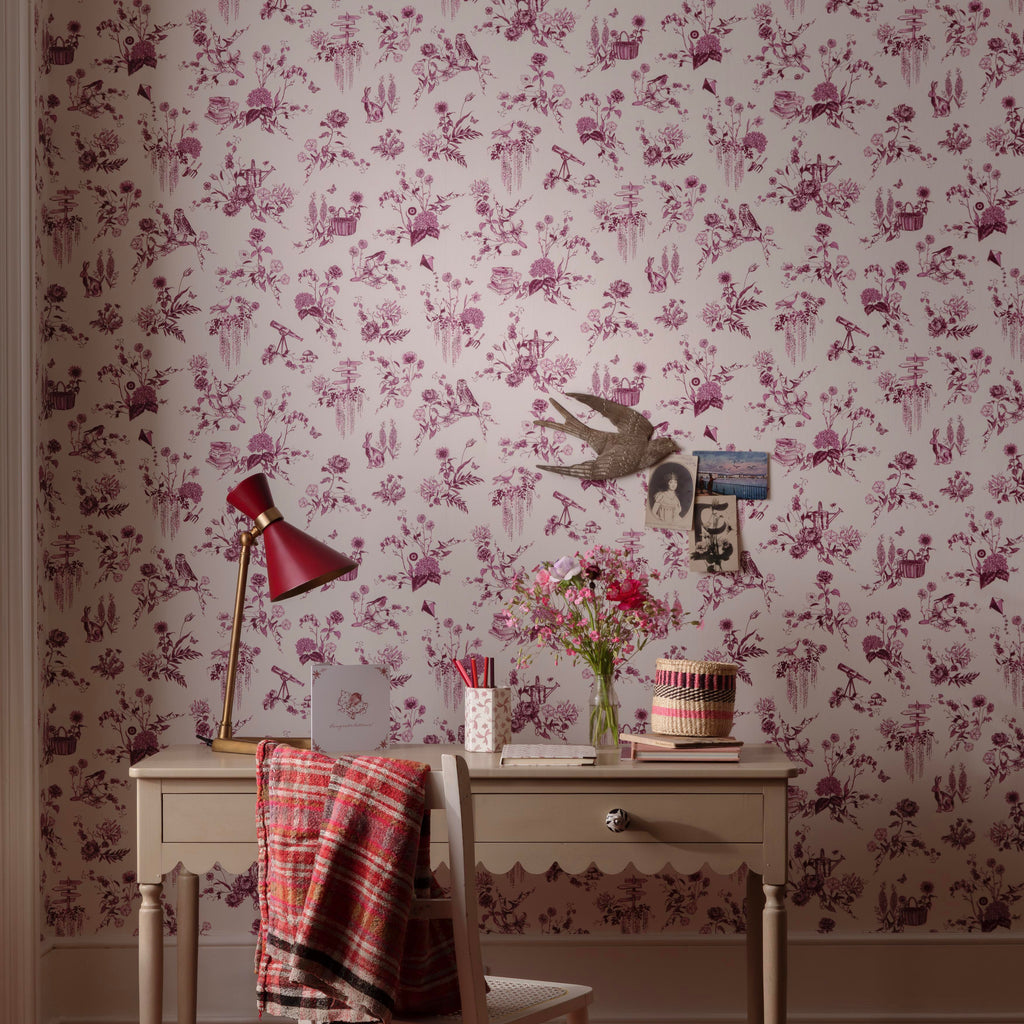 A styled interior scene featuring floral wallpaper with a purple and pink toile-style pattern, a vintage-style desk, a chair with a plaid throw, a red desk lamp, and a striped red-and-white rug.