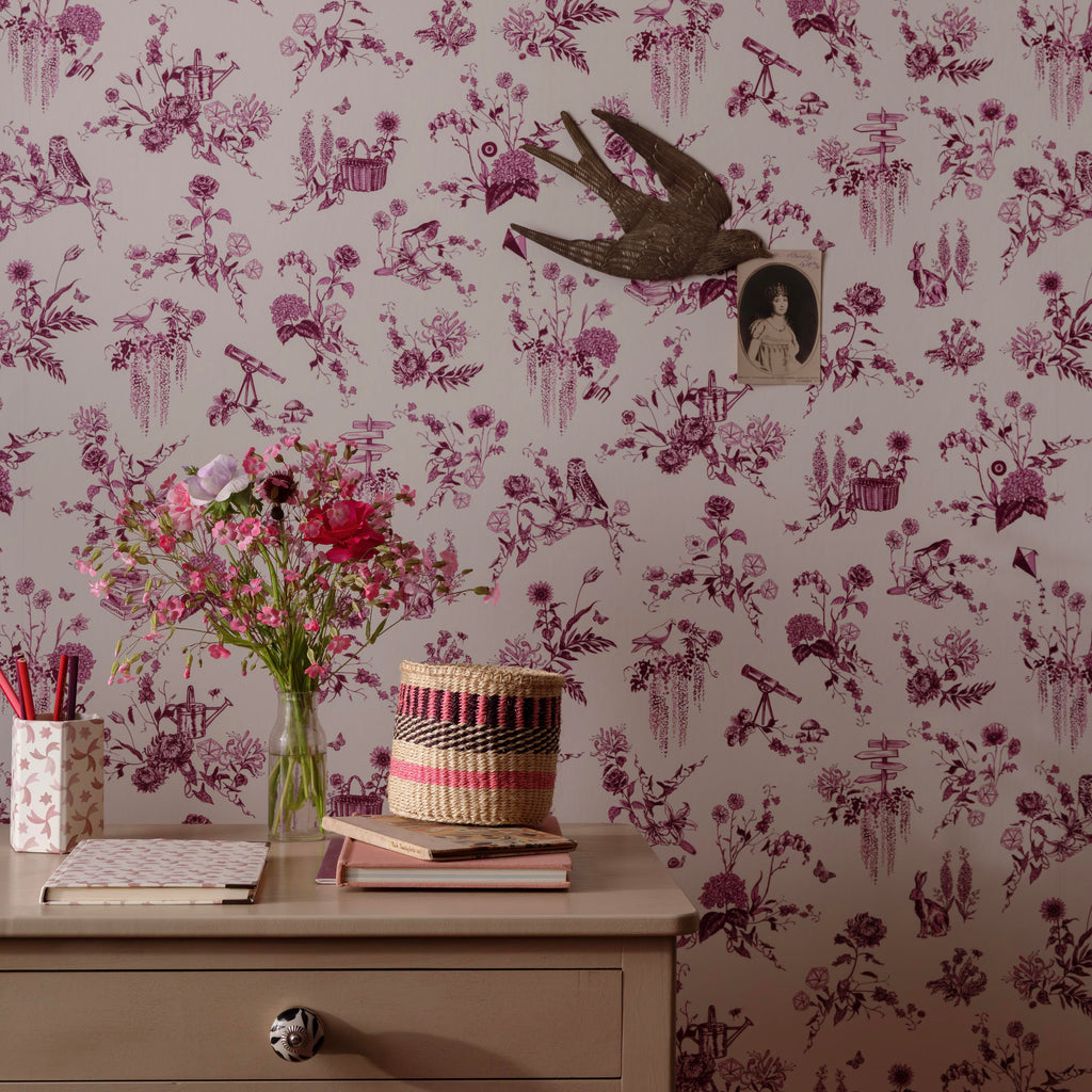 A styled interior scene showing a wall covered in purple toile-style floral wallpaper with birds and floral motifs. A pale cream table at the bottom holds a vase of pink flowers, stacked books, and a small basket, with a small bird-shaped wall decor piece hanging on the wall.