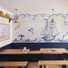 A nautical-themed restaurant interior featuring a large blue and white tiled wall mural with a sailing ship, waves, seagulls, and a lighthouse, above dark blue wainscoting and wooden tables with wooden benches.