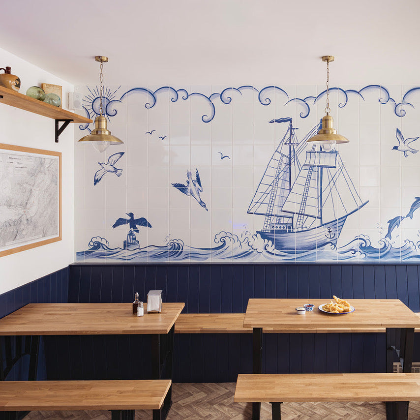 A nautical-themed restaurant interior featuring a large blue and white tiled wall mural with a sailing ship, waves, seagulls, and a lighthouse, above dark blue wainscoting and wooden tables with wooden benches.