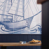 A large blue and white nautical wall mural featuring a detailed sailing ship and waves, mounted above a dark blue paneled wall and a wooden table with food in the foreground.