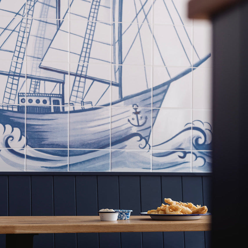 A large blue and white nautical wall mural featuring a detailed sailing ship and waves, mounted above a dark blue paneled wall and a wooden table with food in the foreground.