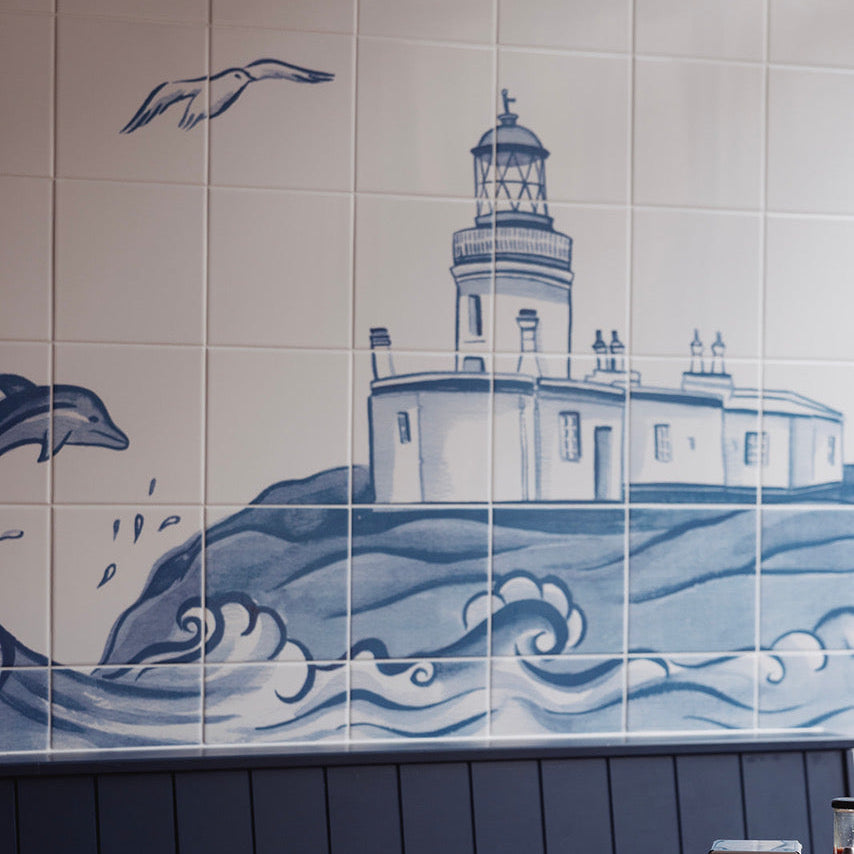 Blue and white tiled wall mural depicting a lighthouse on waves, with a wooden table and navy wainscoting in the foreground.