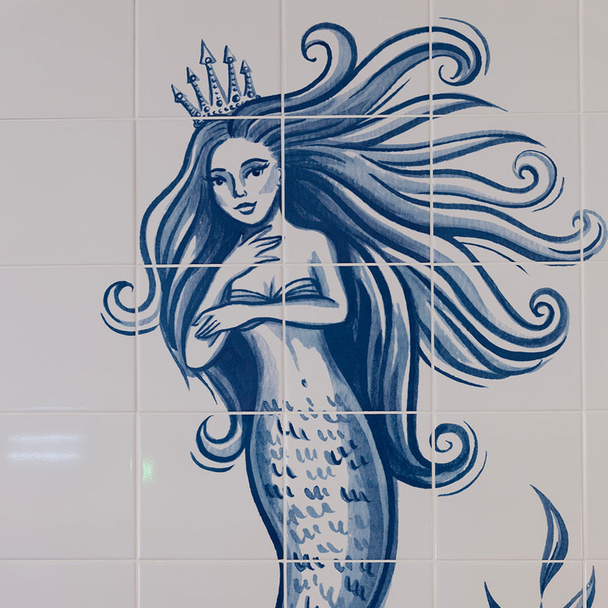 Blue and white ceramic tile mural featuring a mermaid with long flowing hair, a crown, and a scaled tail, with a decorative plant motif in the lower right corner.