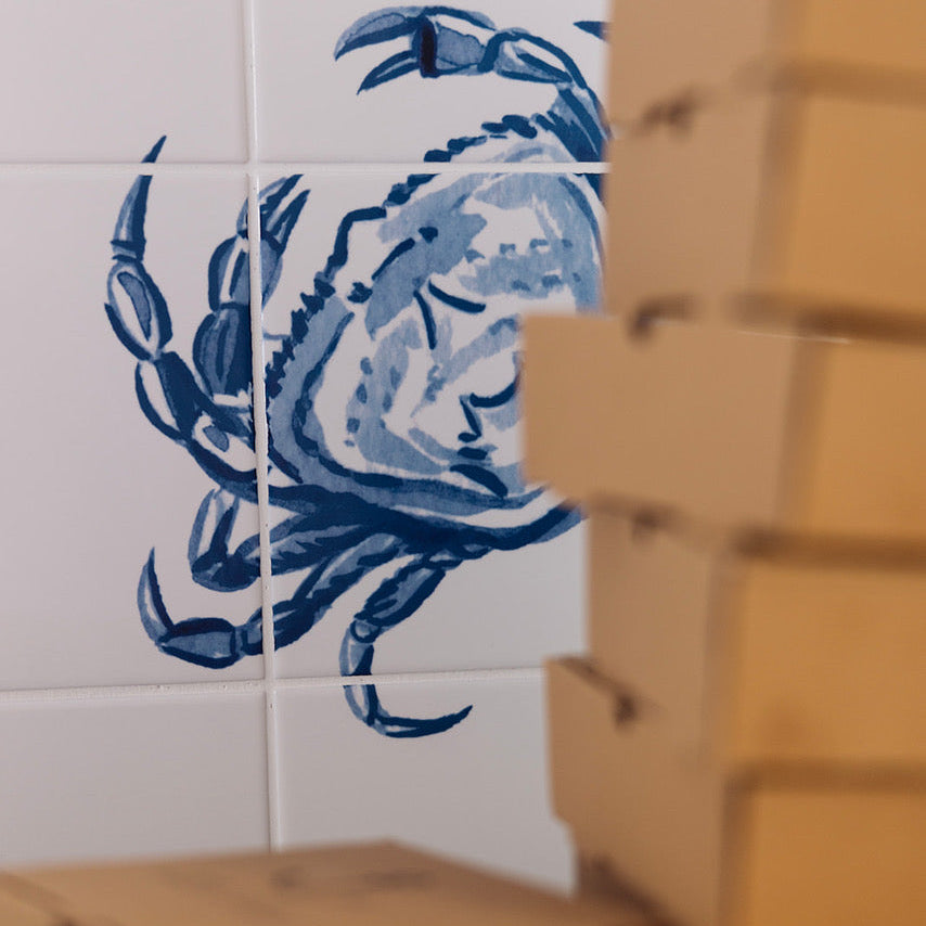 White ceramic tiles featuring a blue hand-painted style crab motif, with stacks of brown cardboard boxes partially obscuring the right side and foreground of the photo.