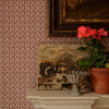 A styled interior corner showing patterned wallpaper in muted pink and brown, a white fireplace mantel, a framed painting, a small landscape artwork, and a potted pink flower.