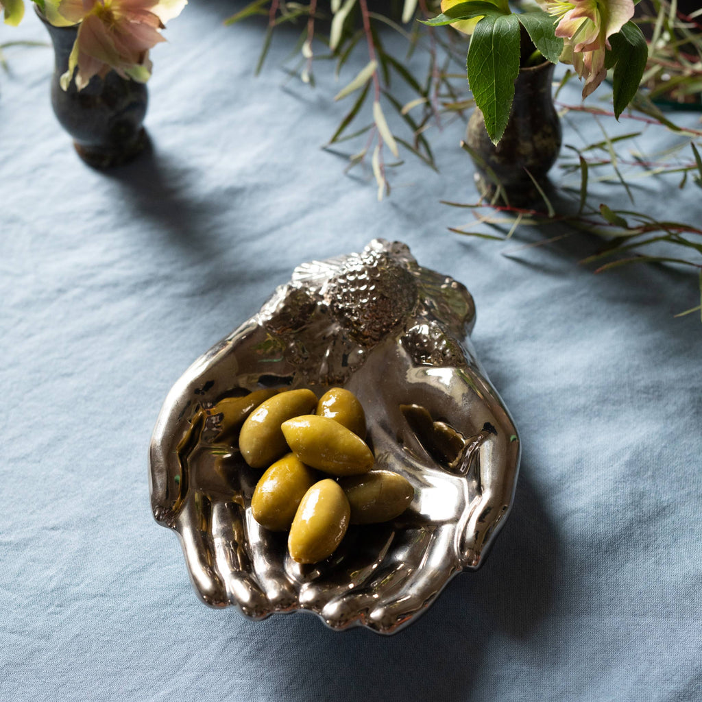 Decorative hand shaped silver dish with olives on a textured blue surface with floral elements.