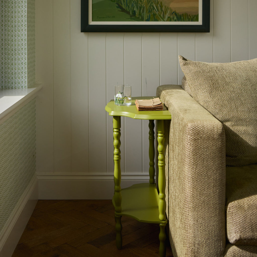 Living room with a framed painting on the wall, a green side table with a hand painted glass, and a beige sofa.