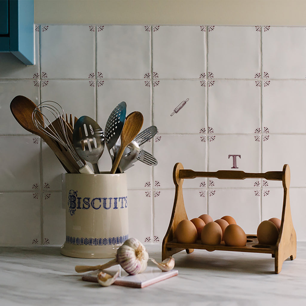 A styled kitchen scene with a cream ceramic biscuit jar filled with wooden and metal utensils, a small wooden egg holder containing brown eggs, garlic, and a notebook on a light marble-like surface, set against white patterned delft tiles decorated with a letter T and rolling pin illustration.