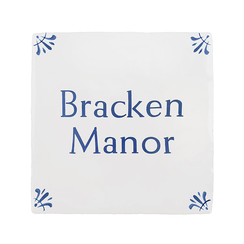 A square white decorative tile or plaque with the words 'Bracken Manor' printed in blue serif text, with small blue floral corner motifs, shown on a plain white background.
