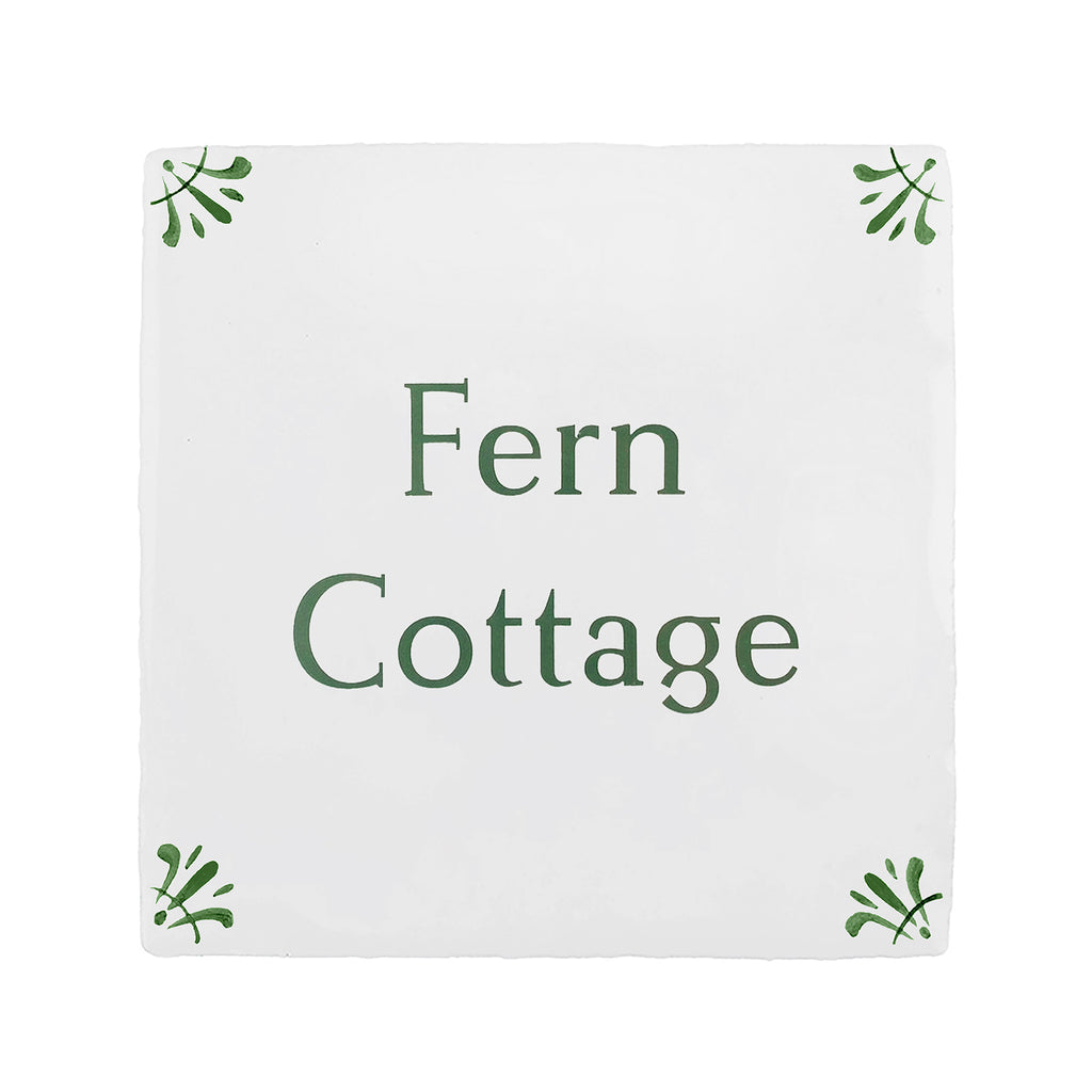 A square white decorative tile or sign with the words 'Fern Cottage' printed in dark green serif text, with small green leaf motifs in each corner, shown on a plain white background.