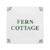 A square white tile-style sign with the words 'FERN COTTAGE' in dark green serif lettering, decorated with small green leaf motifs in each corner, shown on a plain white background.