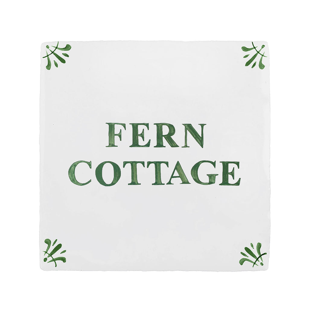 A square white tile-style sign with the words 'FERN COTTAGE' in dark green serif lettering, decorated with small green leaf motifs in each corner, shown on a plain white background.