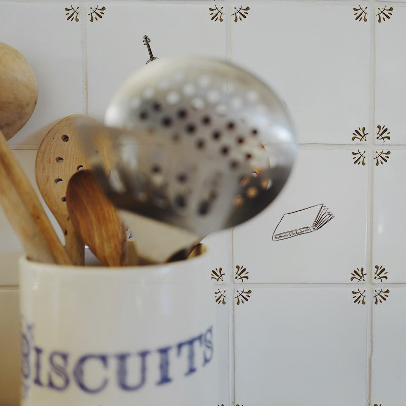 A close-up of kitchen utensils in a white ceramic container labeled 'BISCUITS' against a white tiled wall with small decorative motifs.