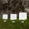 Three square white tiles with blue floral designs are displayed on grass in front of a stone wall, labeled 10cm, 12.5cm, and 15cm in size.
