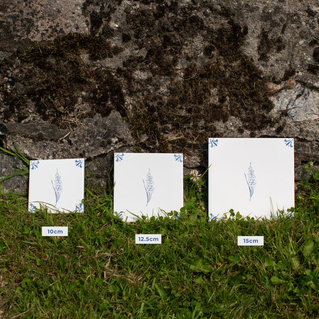 Three square white tiles with blue floral designs are displayed on grass in front of a stone wall, labeled 10cm, 12.5cm, and 15cm in size.