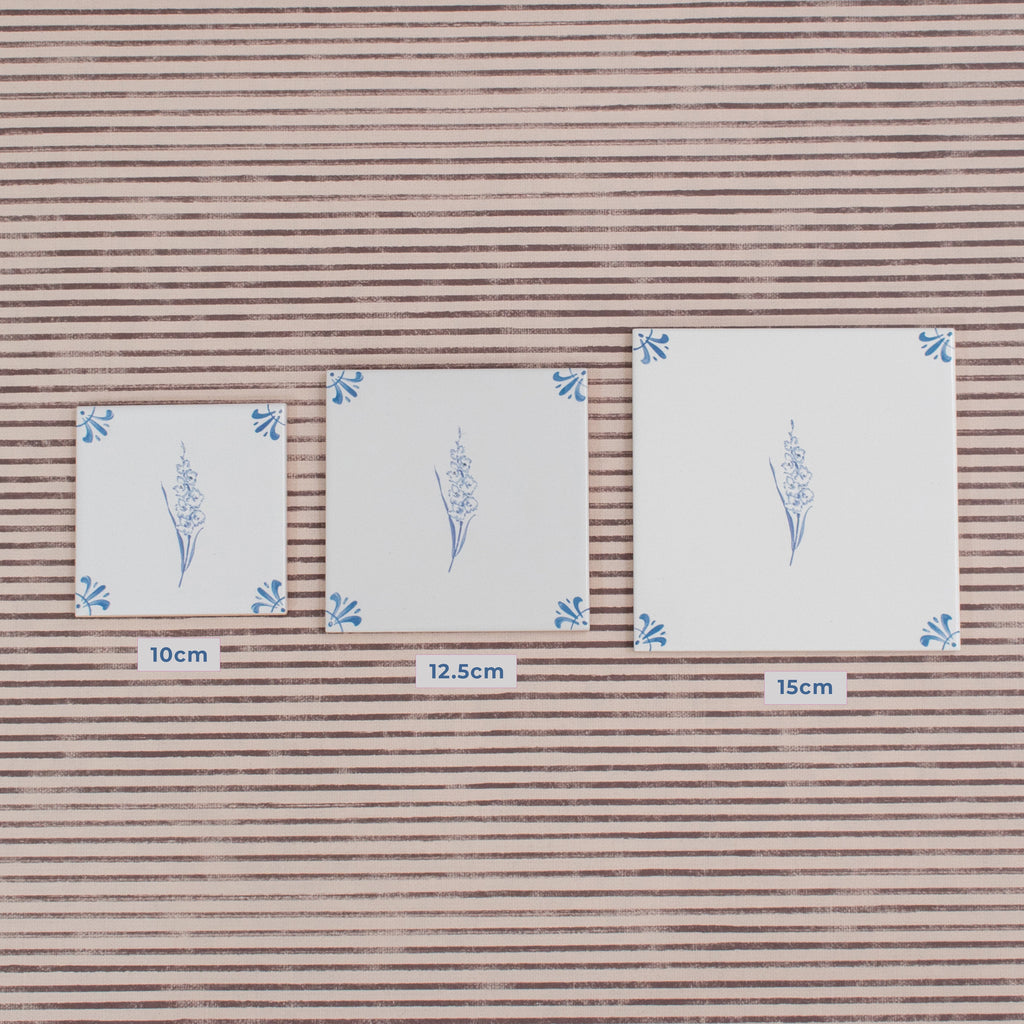 Three square white tiles with blue floral designs are shown on a background of striped wallpaper, comparing the size of a 10cm, 12.5 and 15cm tile