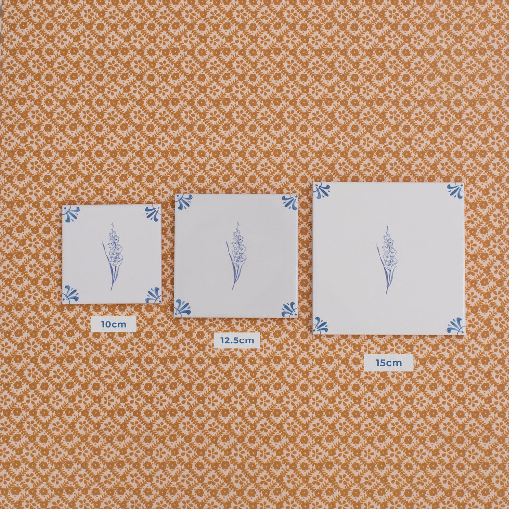 Three square white ceramic tiles with blue floral designs are laid out on a patterned orange wallpaper, comparing the sizes of 10cm, 12.5 and 15cm tiles